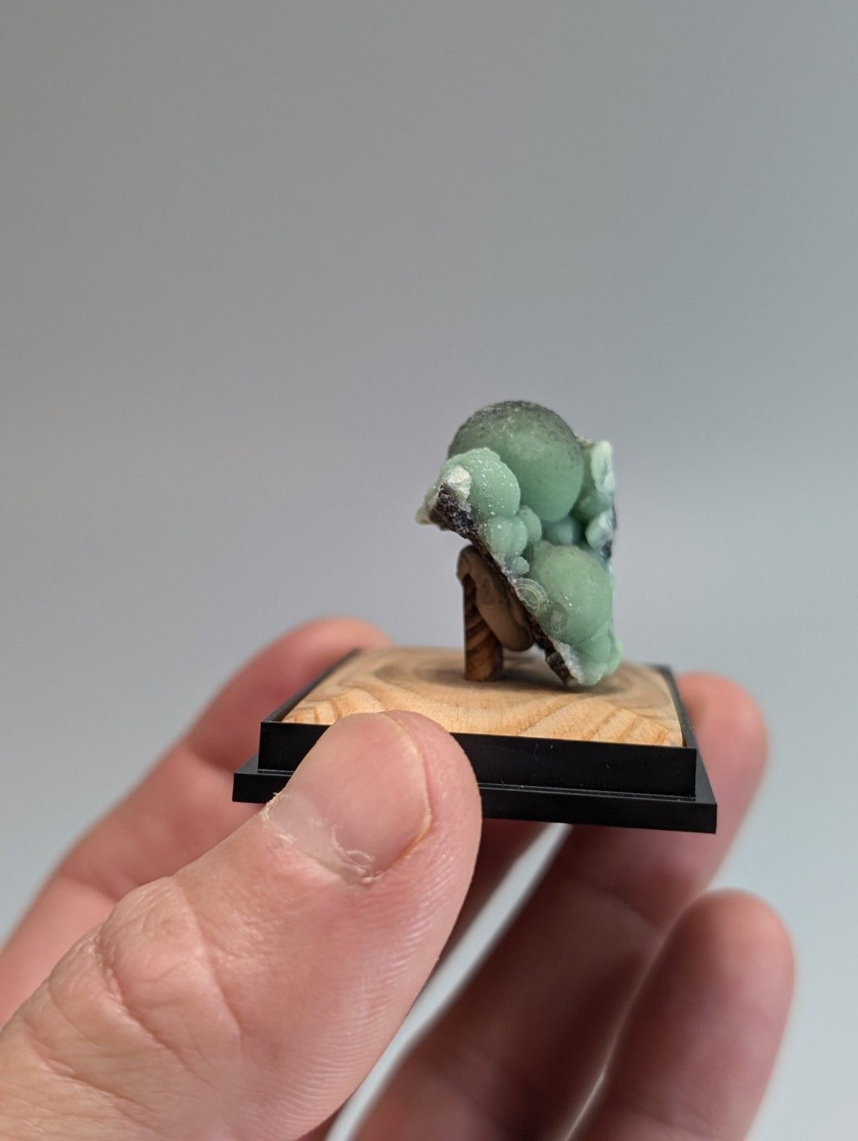 A miniature green jade-like mineral specimen with a textured, bubbly surface mounted on a wooden base.