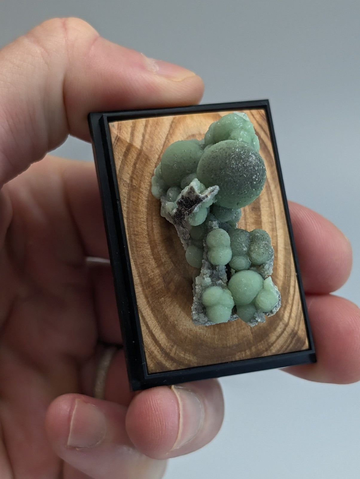 A framed green spherical mineral specimen with a natural wood background and black frame.
