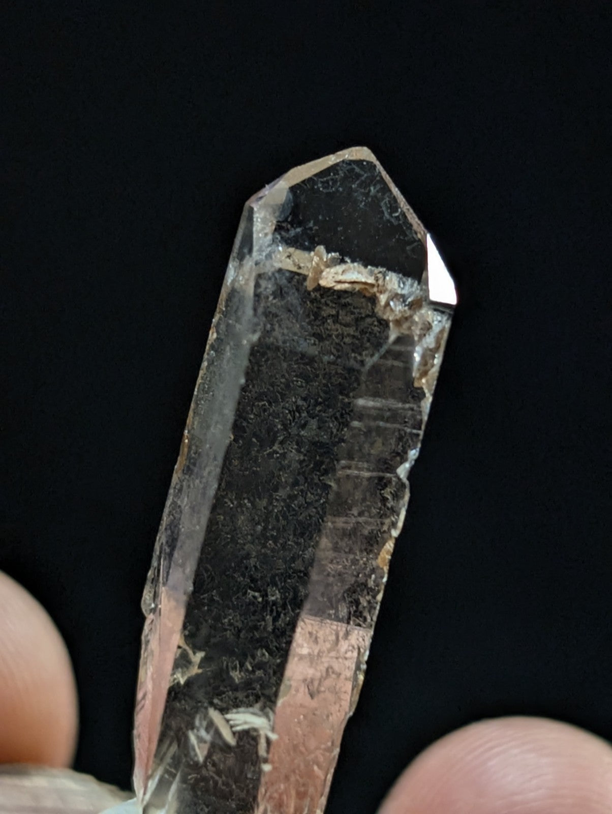 A clear quartz crystal with a pointed tip and visible internal inclusions, held between fingers against a dark background.