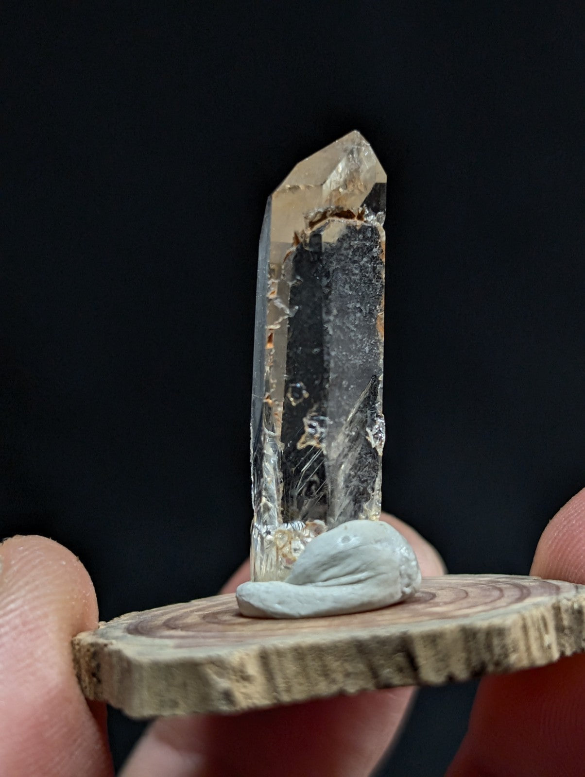 A clear, faceted quartz crystal with a natural, slightly rough texture and subtle internal inclusions stands upright on a small wooden slice.