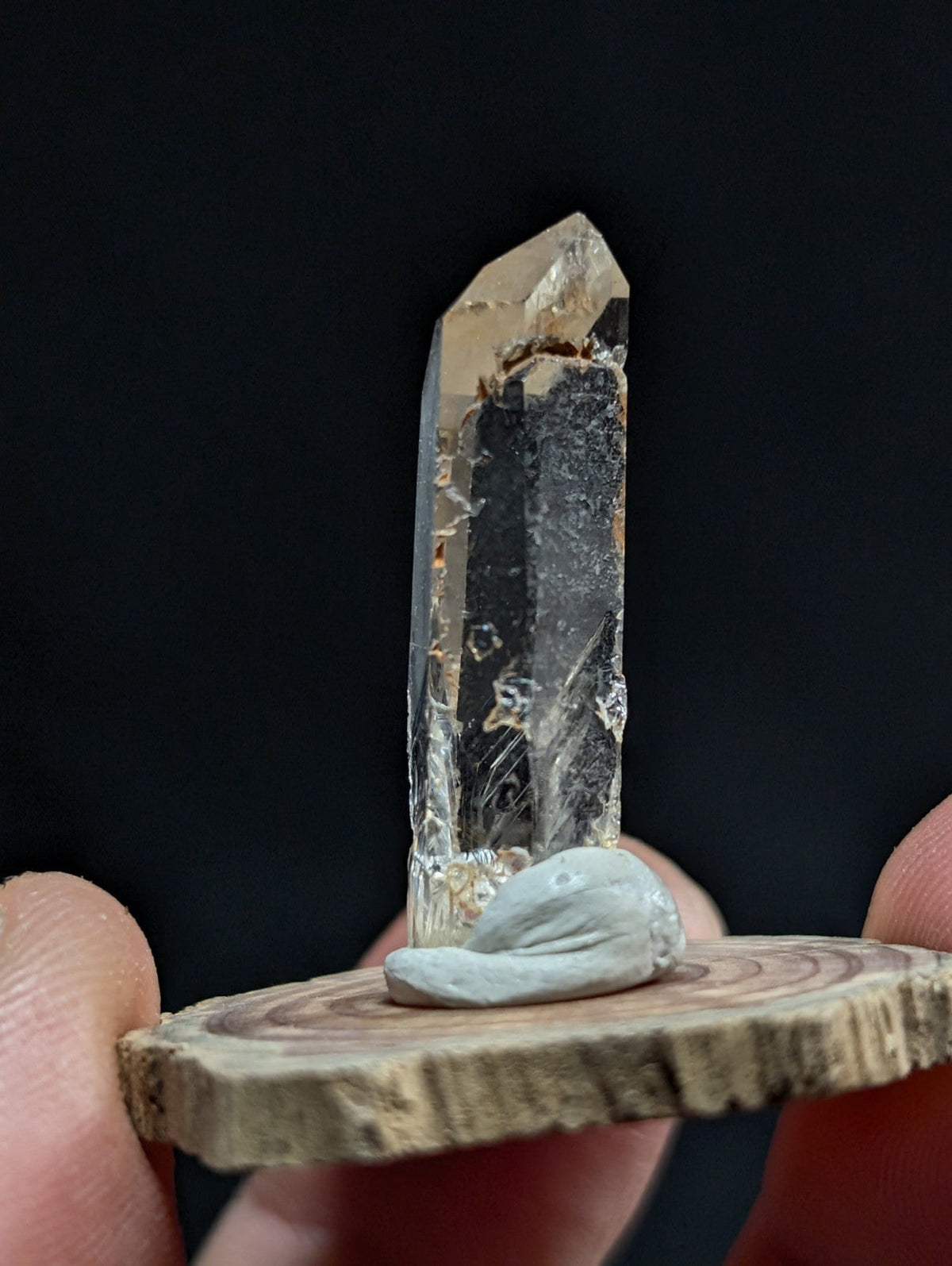 A clear, faceted quartz crystal with a natural, slightly rough texture and subtle internal inclusions stands upright on a small wooden slice.