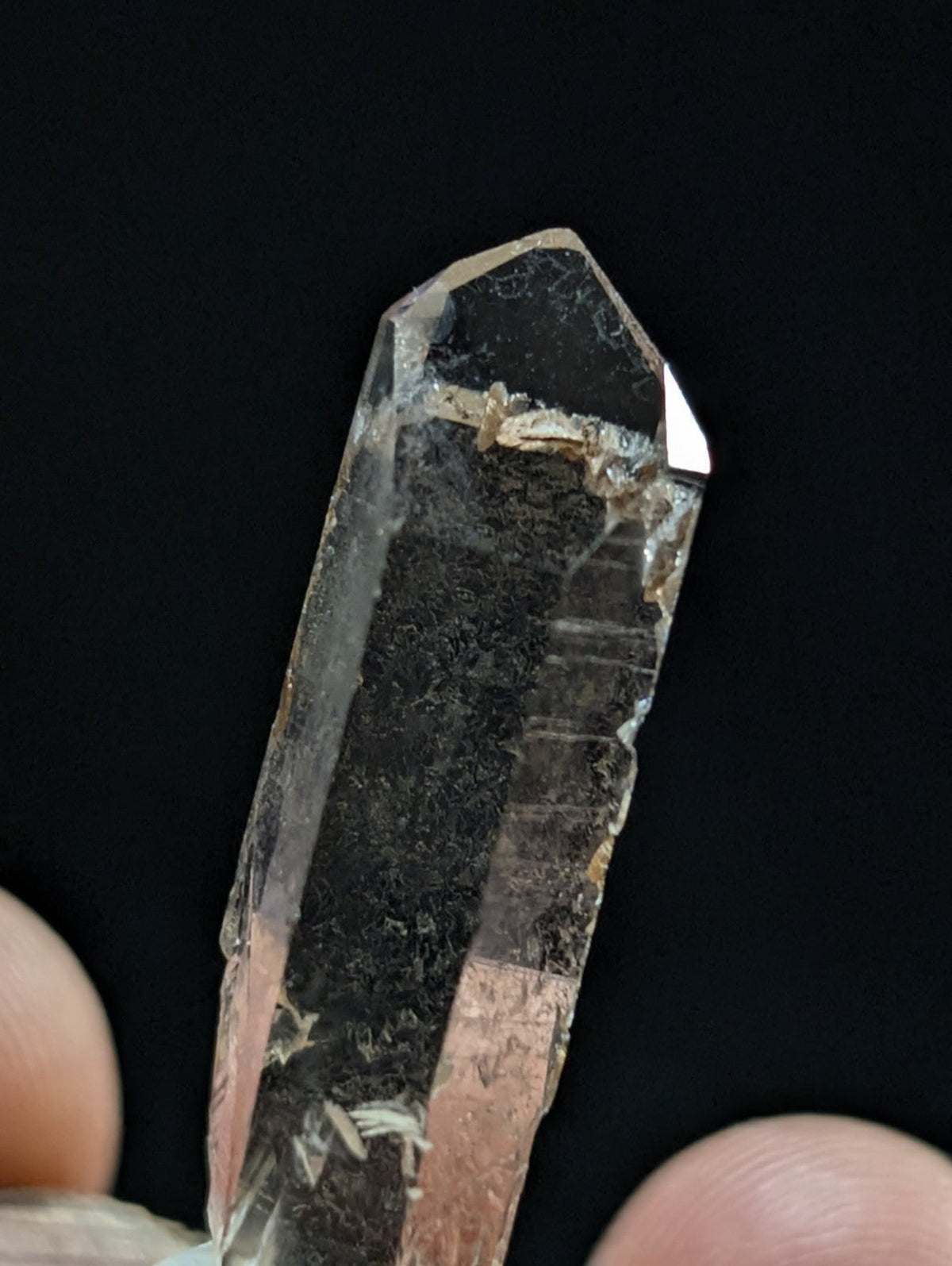 A clear quartz crystal with a pointed tip and visible internal inclusions, held between fingers against a dark background.
