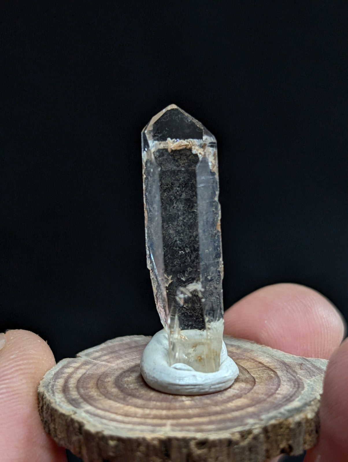 A clear quartz crystal with a pointed tip and natural inclusions, mounted on a circular white base atop a rustic wooden slice.