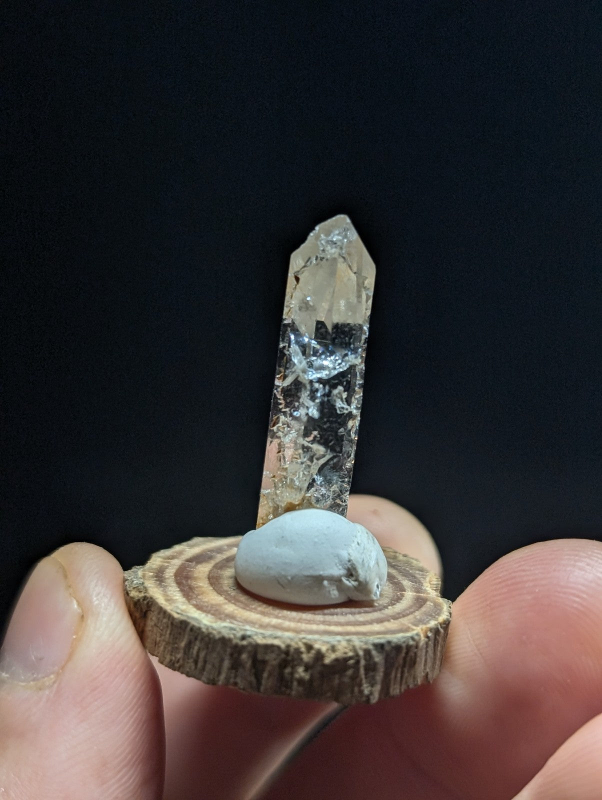 A clear quartz crystal with a pointed tip and internal inclusions, mounted on a circular wooden slice base.