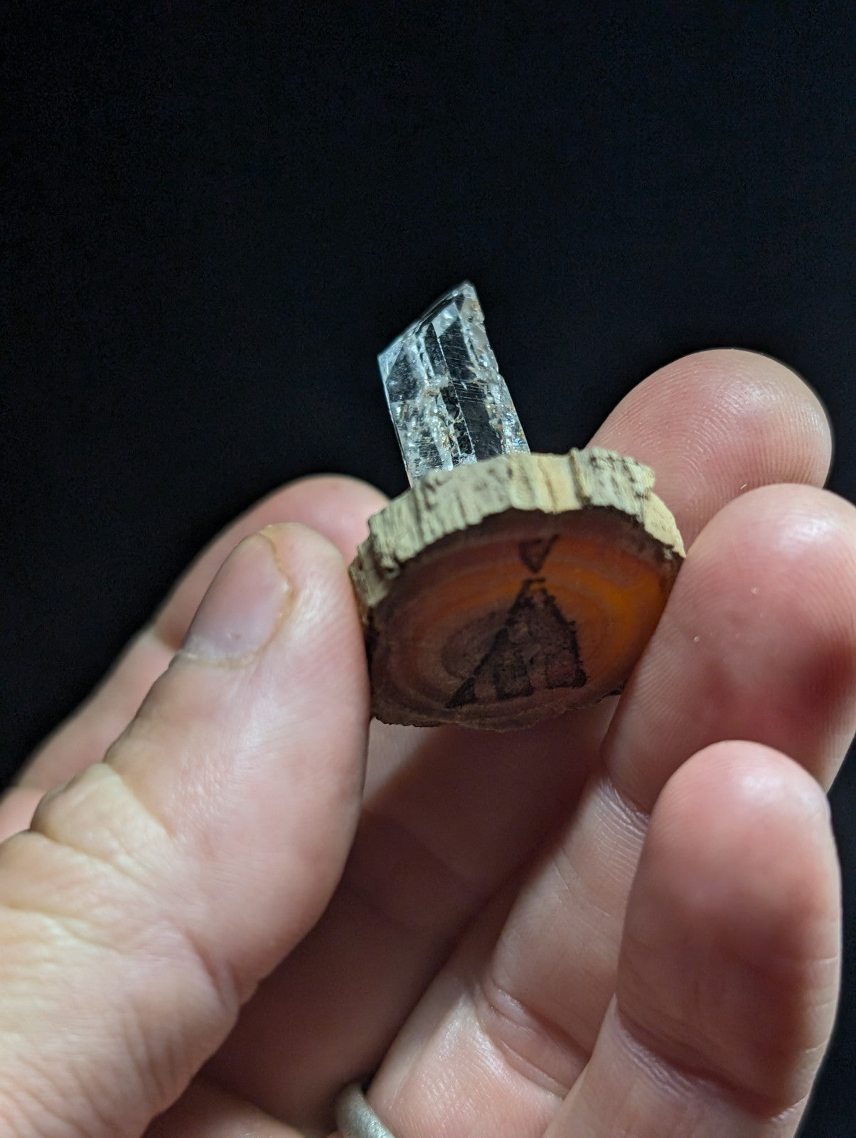 A clear, faceted quartz crystal mounted on a circular slice of wood with an engraved teepee design.