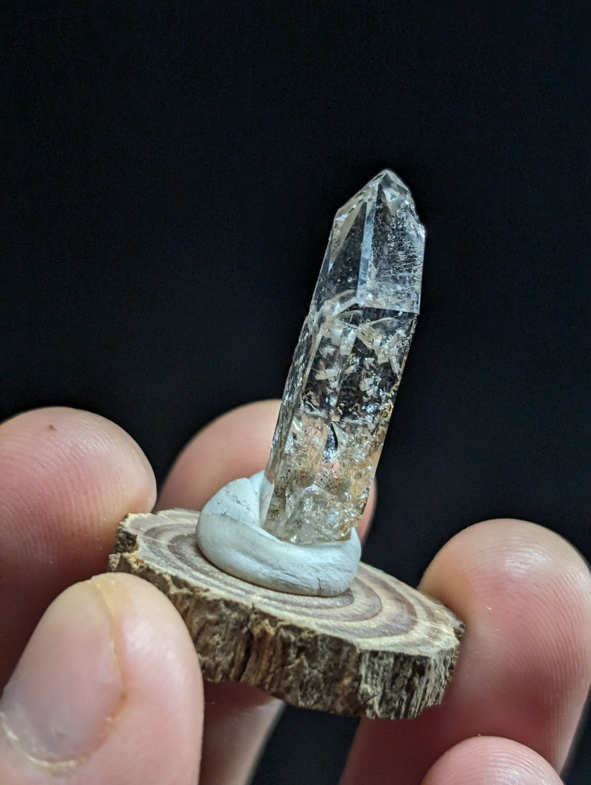 A clear, faceted quartz crystal stands upright on a circular wooden slice base.