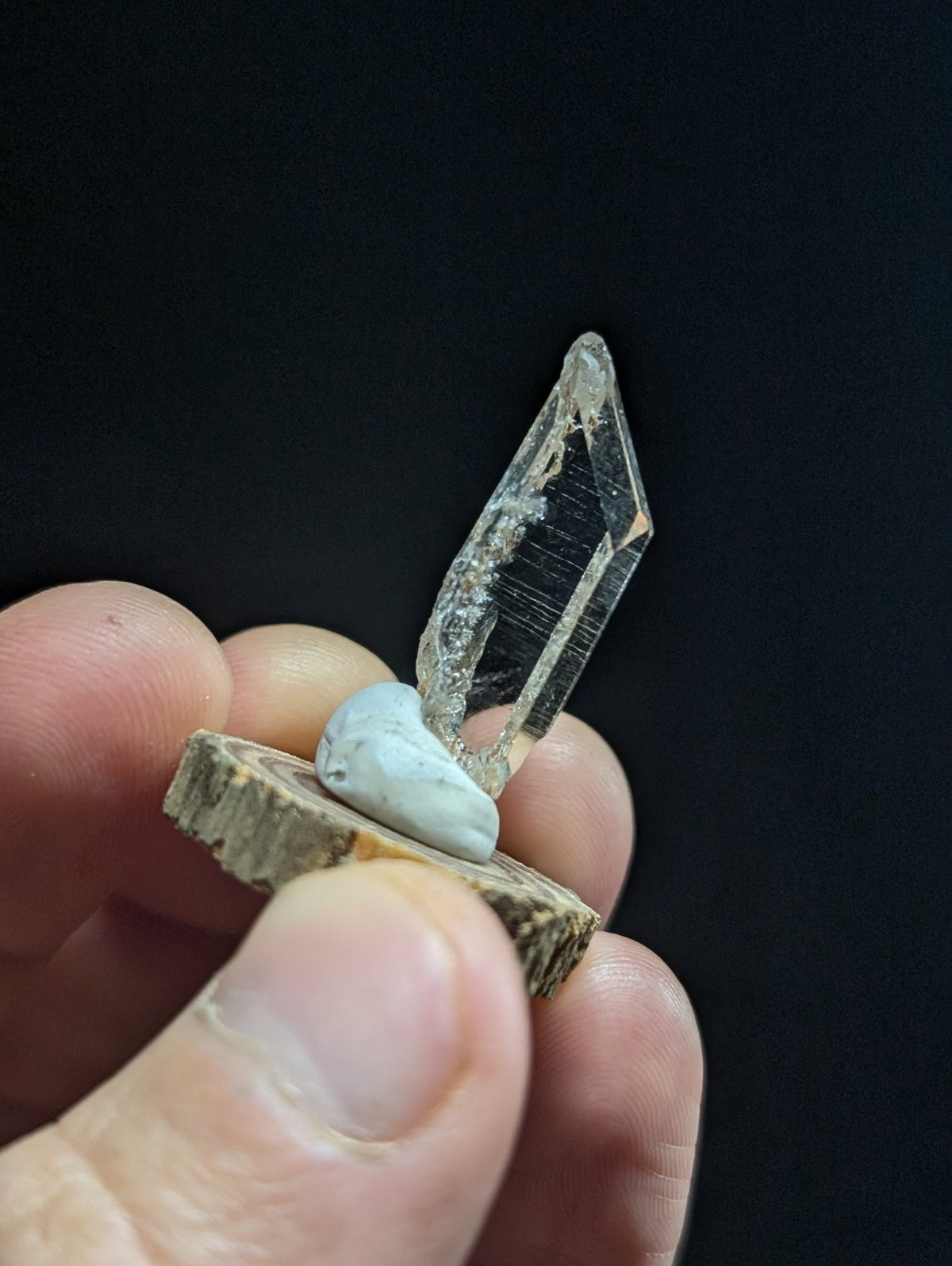 A clear, faceted quartz crystal stands upright on a small, light blue stone atop a rustic wooden slice.