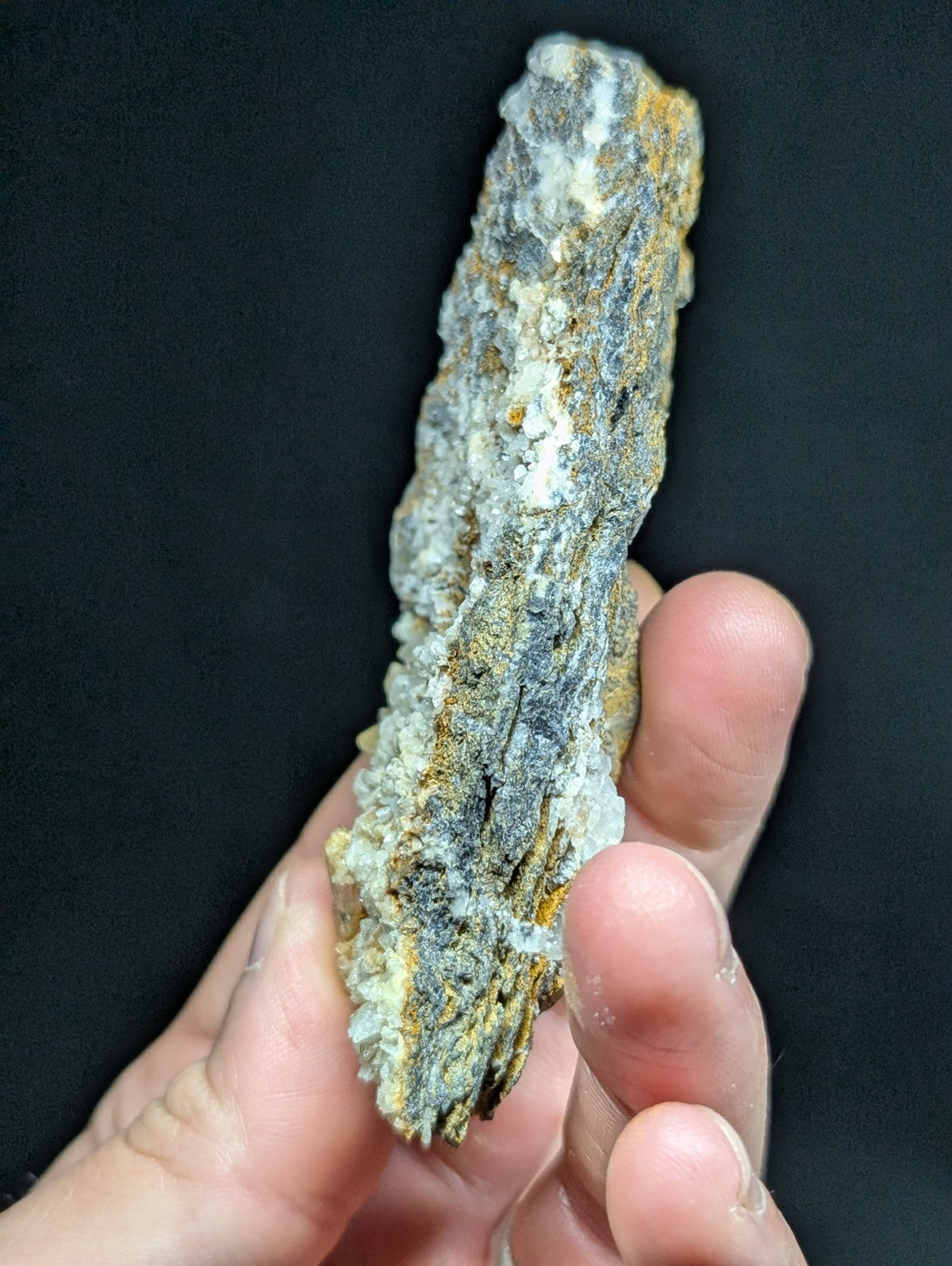 A textured, elongated mineral specimen with a mottled surface of gray, white, and yellowish hues.