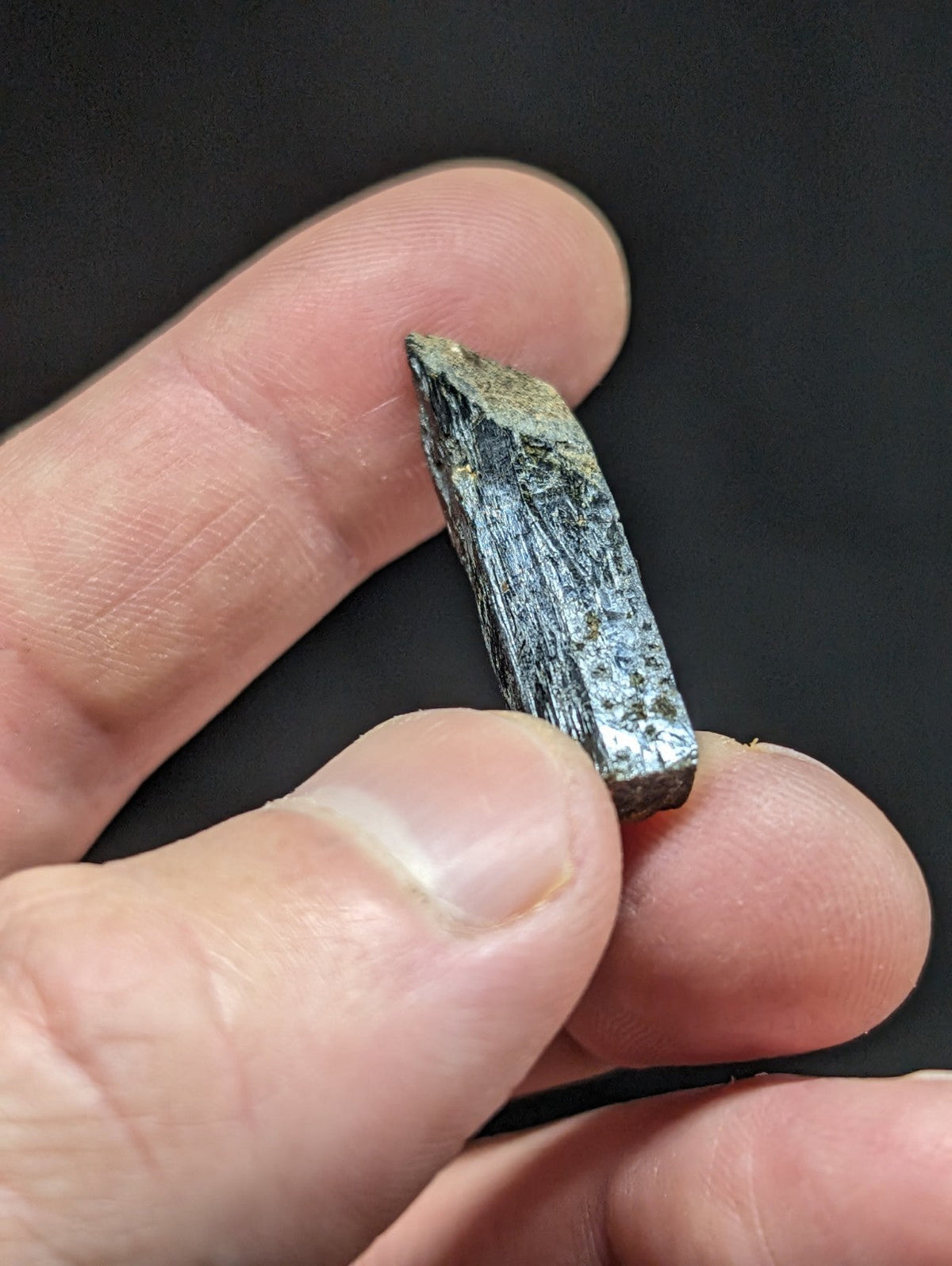 A dark gray, elongated mineral crystal with a rough, faceted surface and subtle greenish veining.