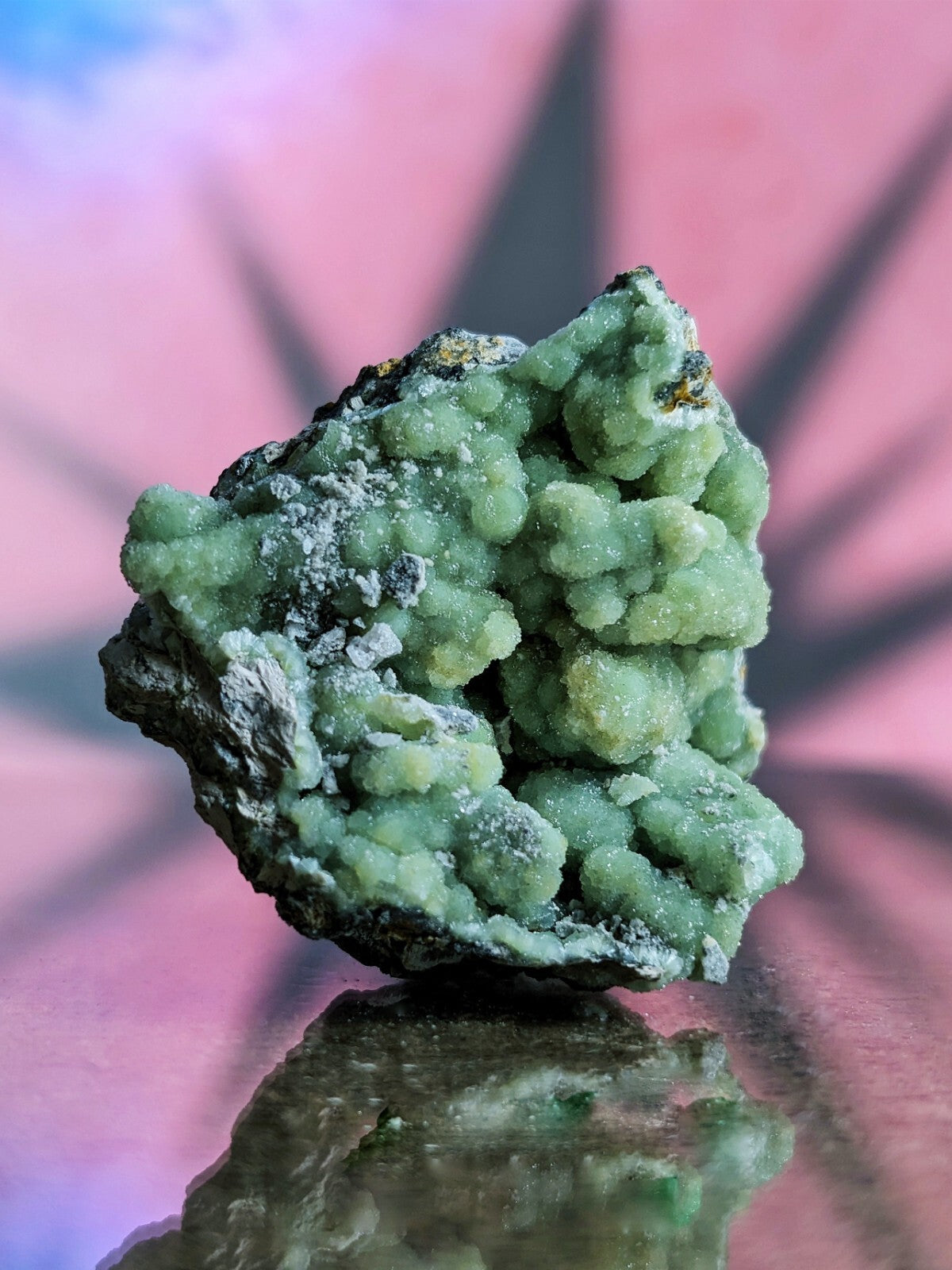 A green, crystalline mineral specimen with a rough, textured surface and patches of dark matrix.