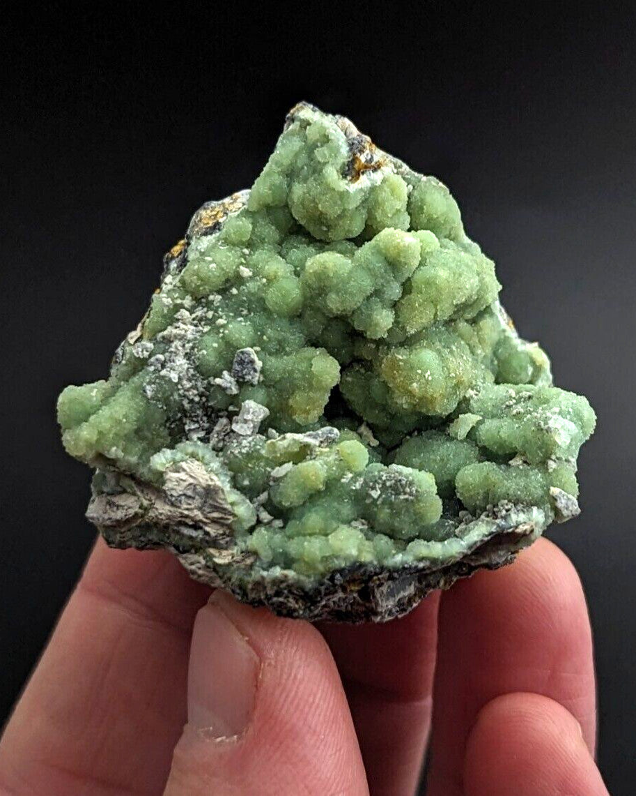 A hand holds a rough, irregularly shaped mineral specimen featuring vibrant green crystalline formations with some white and gray matrix material.