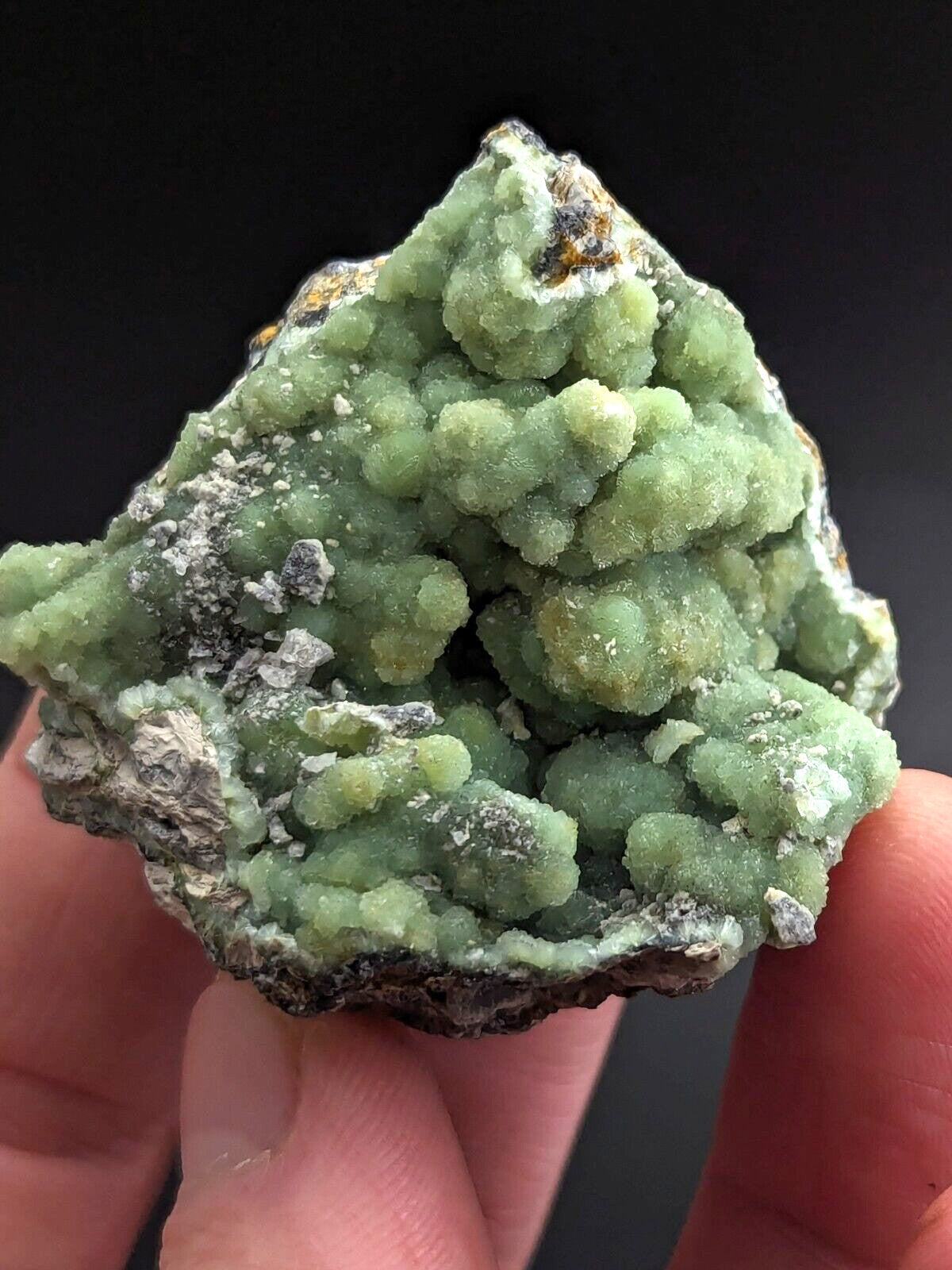 A vibrant green, crystalline mineral specimen with a rough, textured surface and scattered white and dark patches.
