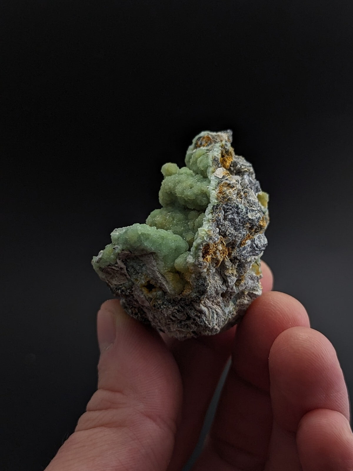 A hand holds a natural mineral specimen featuring vibrant green crystalline formations growing from a rough, grayish matrix with patches of yellow and white.