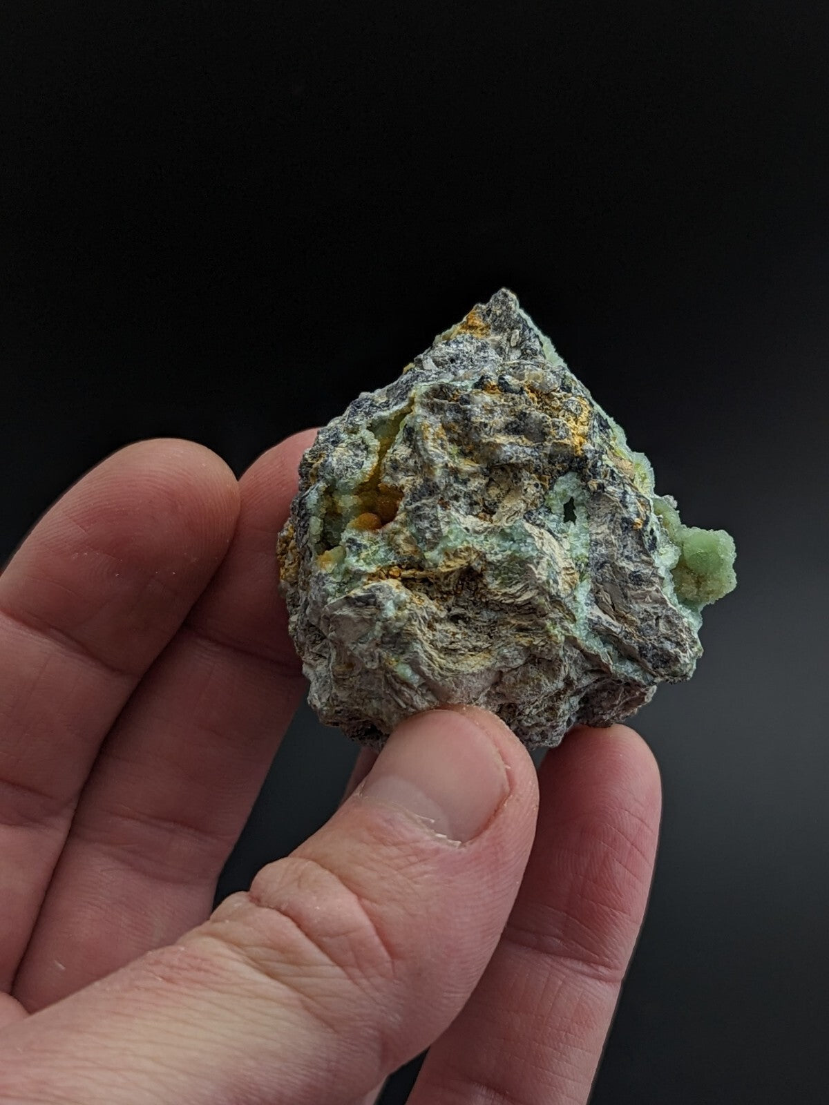 A rough, irregularly shaped mineral specimen with a mottled gray surface accented by patches of green and yellow.