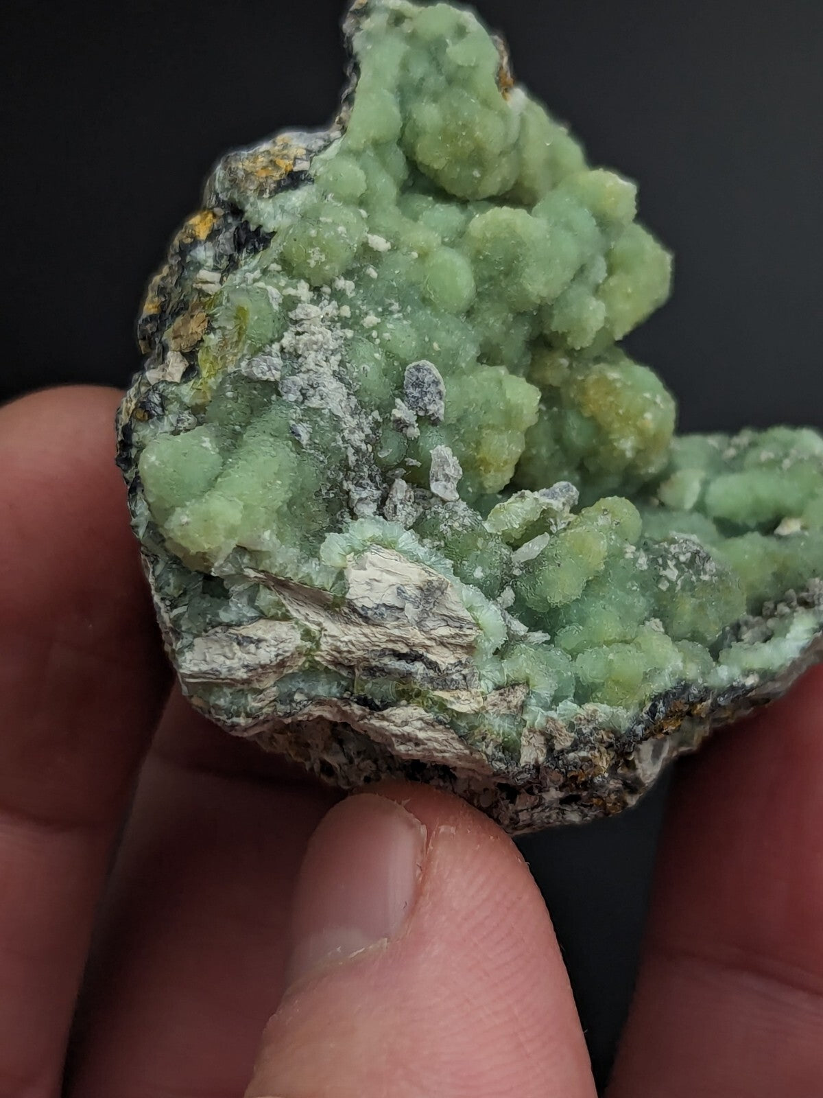 A hand holds a green, crystalline mineral specimen with clustered, rounded formations and patches of white and yellowish matrix.