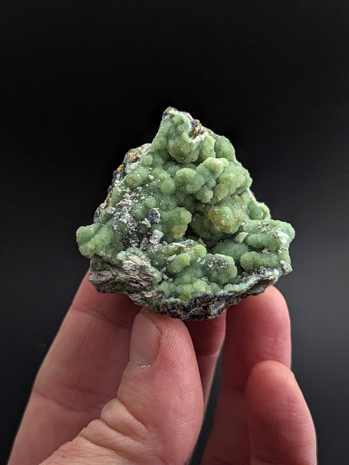 A hand holds a vibrant green mineral specimen with clustered, bumpy formations and patches of white crystalline material against a dark background.