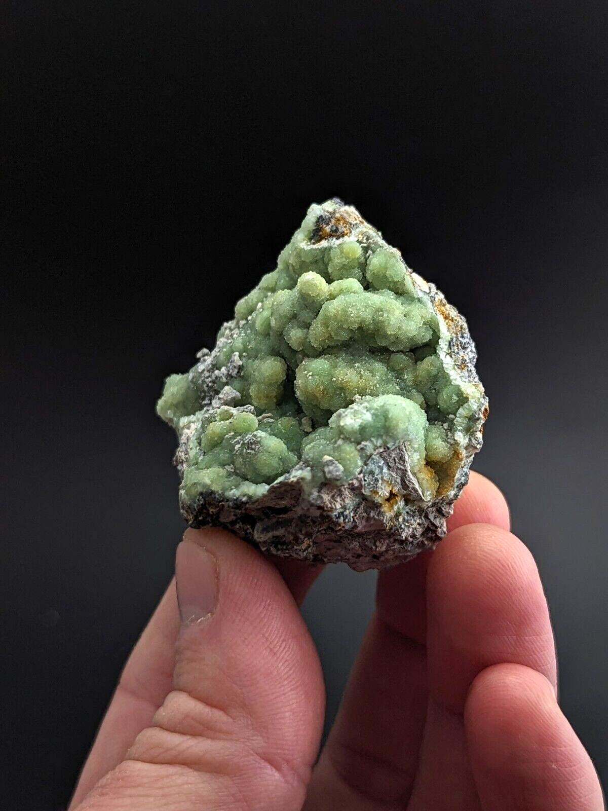 A hand holds a small, irregularly shaped mineral specimen featuring vibrant green crystalline formations clustered atop a darker, rough-textured base with hints of yellow and white.