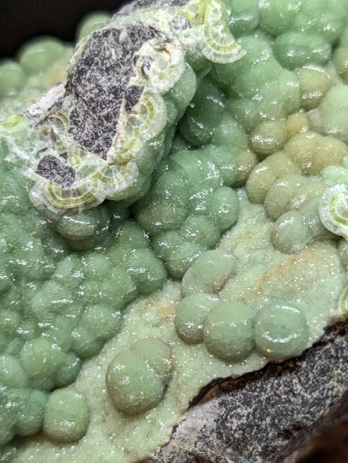 A cluster of translucent green, bead-like mineral formations with a rough-textured surface and some darker grayish patches.