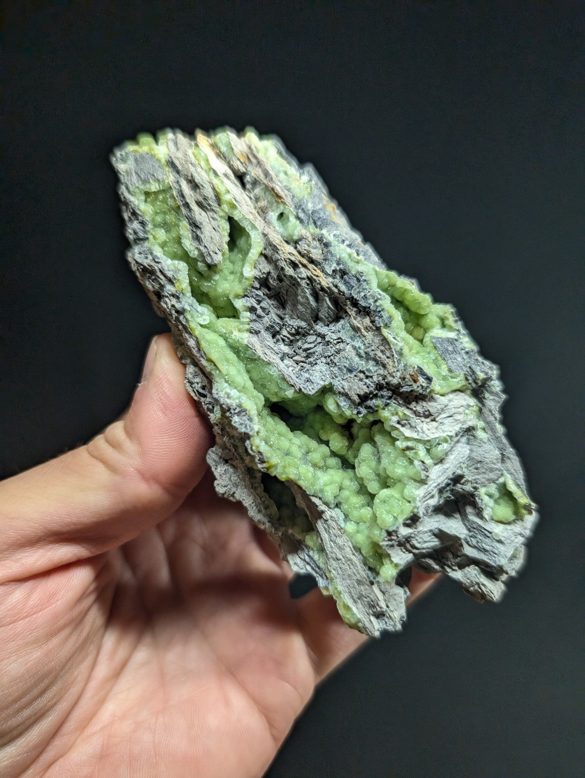 A hand holds a rough, irregularly shaped mineral specimen featuring vibrant green crystalline formations growing from a darker gray matrix.