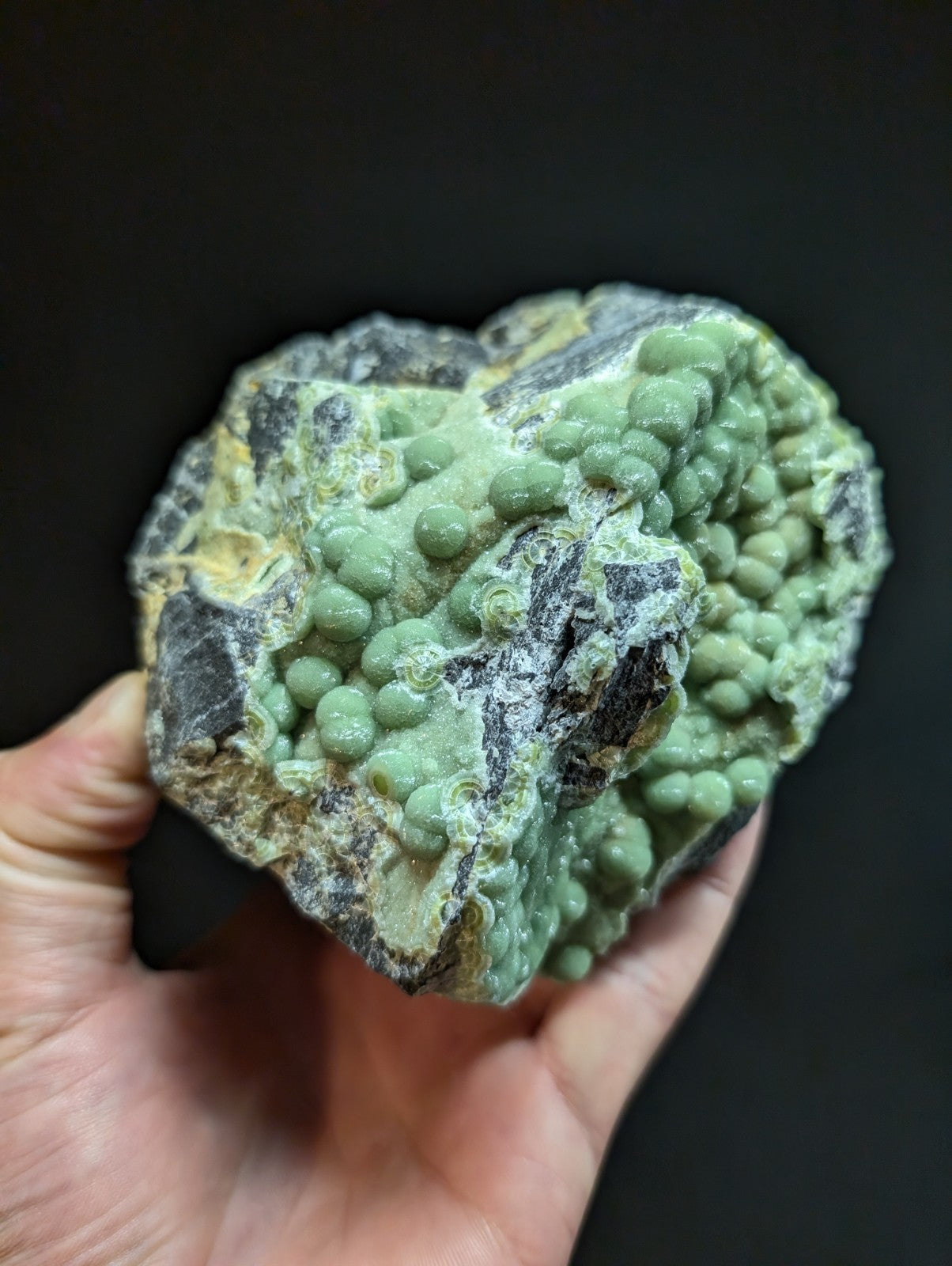 A hand holds a rough, irregularly shaped mineral specimen featuring clusters of small, rounded green nodules embedded in a darker gray and yellowish matrix.