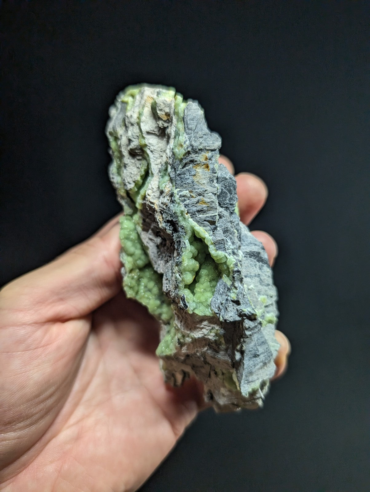 A hand holds a rough, irregularly shaped mineral specimen featuring patches of vibrant green and grayish matrix with visible crystalline textures.
