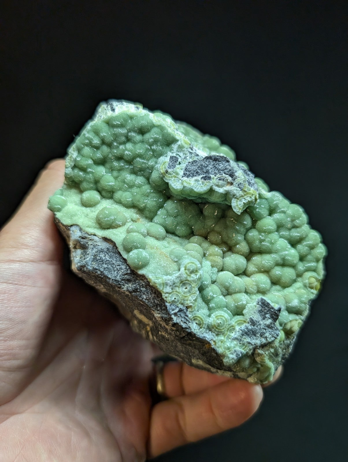 A hand holds a green, bumpy mineral specimen with rounded nodules and dark gray patches.