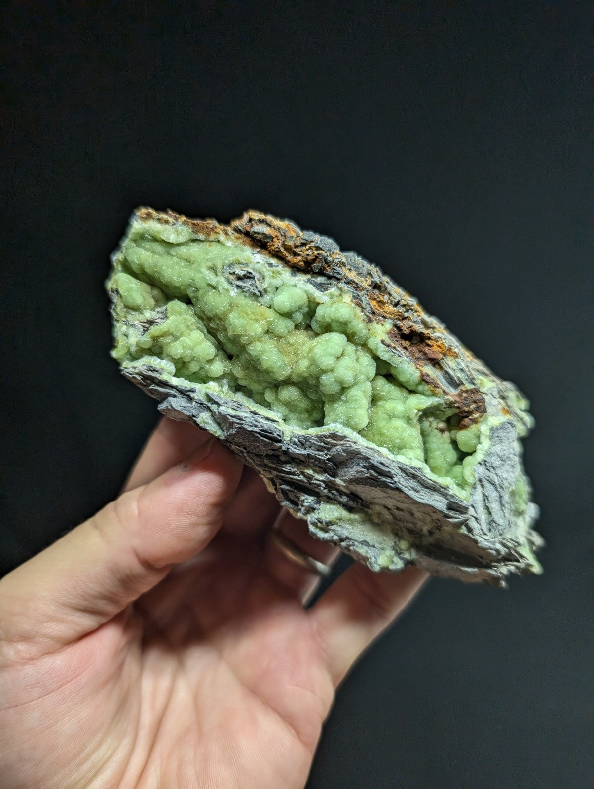 A hand holds a striking green mineral specimen with clustered, bumpy formations against a dark background.