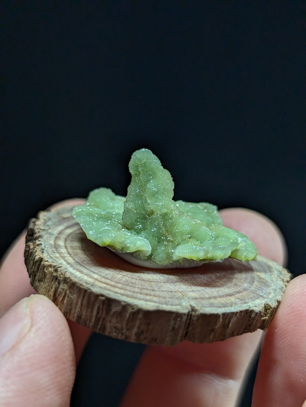 A translucent green crystal formation with a rough, jagged surface sits atop a circular wooden slice.