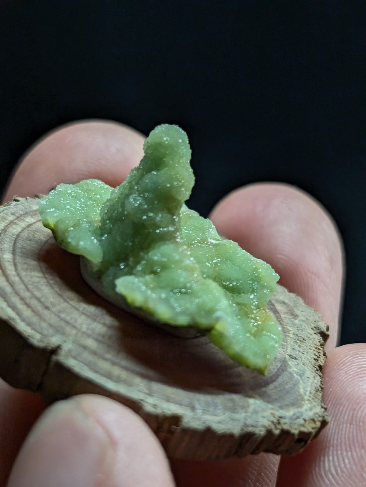 A pale green, translucent mineral specimen with a rough, crystalline texture rests on a circular wooden slice.