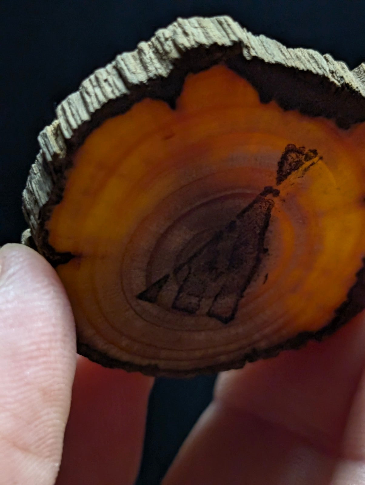 A hand holds a circular slice of wood with a natural, rough bark edge and warm orange-brown interior grain, featuring an engraved dark triangular design.