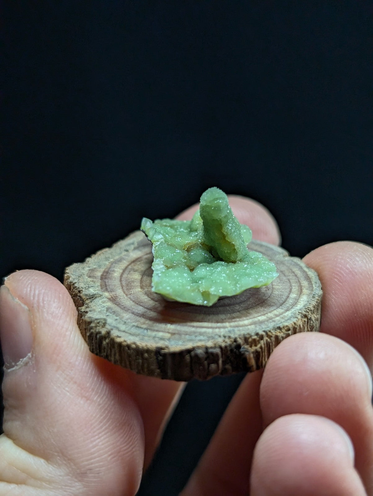 A translucent green crystal formation with a rough, jagged surface sits atop a circular wooden slice.