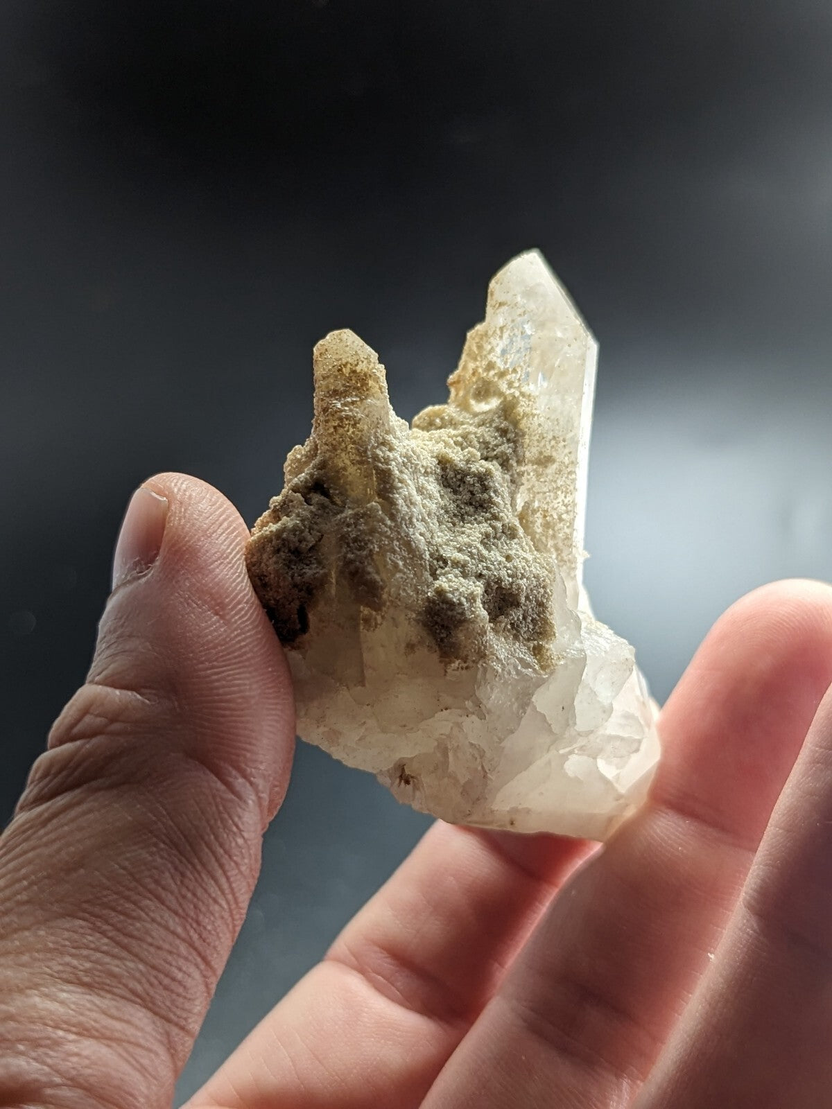 A translucent white quartz crystal with a rough, earthy greenish-brown matrix clinging to its base.