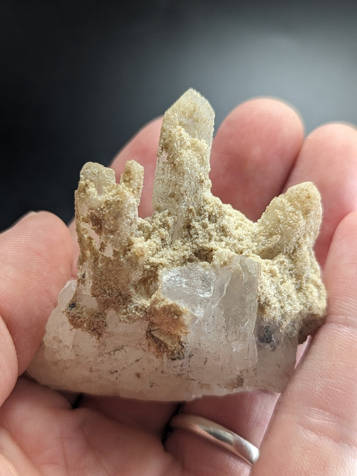 A hand holds a translucent quartz crystal cluster with clear, pointed formations emerging from a rough, earthy matrix.