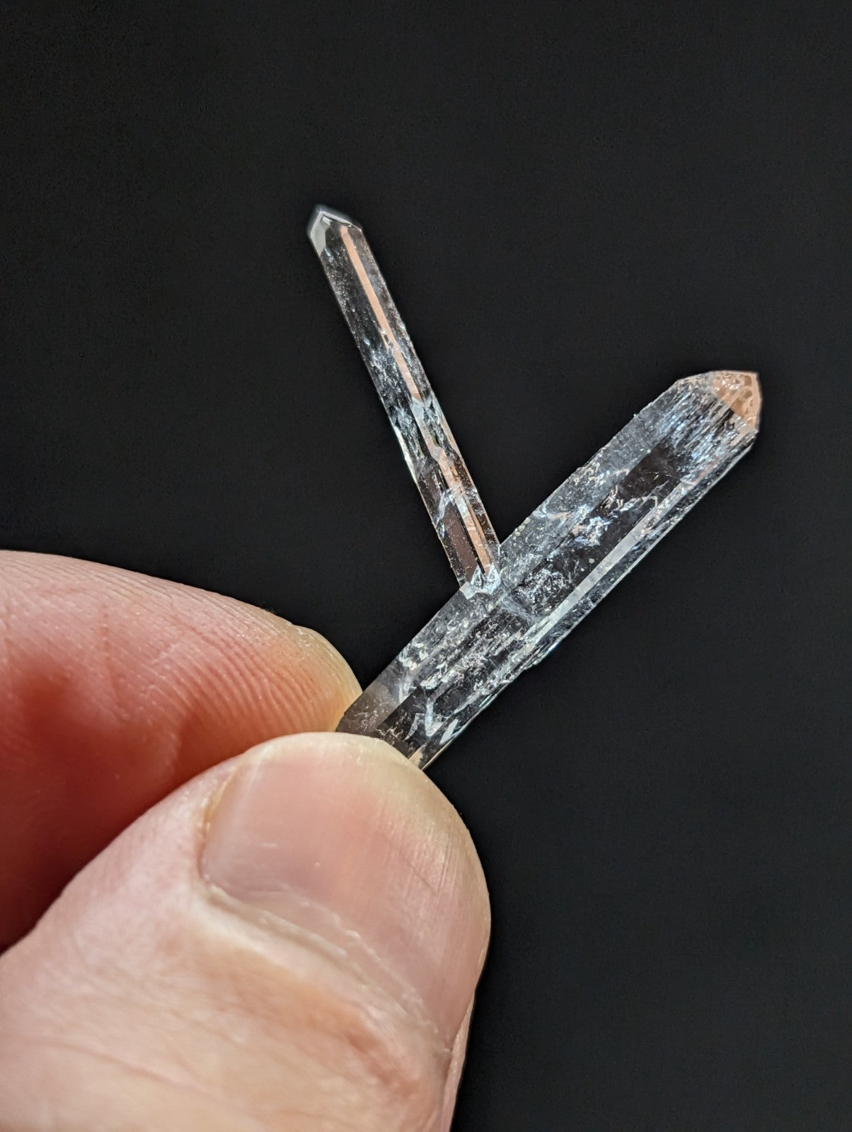 Two translucent quartz crystals, one longer and slightly wider with a pointed tip showing faint brownish discoloration at its base, held between fingers against a dark background.