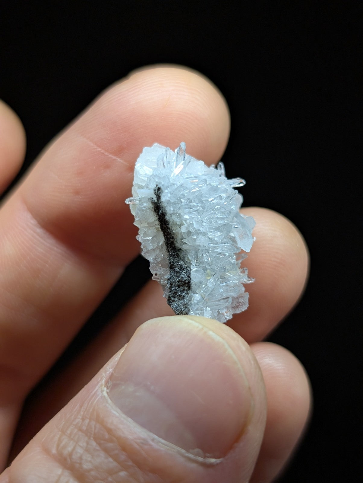 A translucent, crystalline mineral specimen with a delicate, feathery texture and a central dark streak running through it.