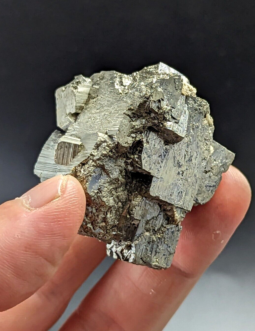 A hand holds a rough, metallic gray pyrite crystal cluster with sharp, angular facets and subtle golden highlights.