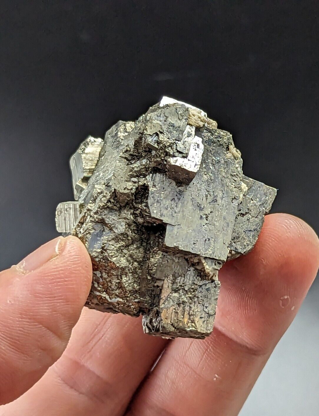 A hand holds a rough, irregularly shaped specimen of pyrite with metallic golden-yellow facets and darker grayish-brown matrix.