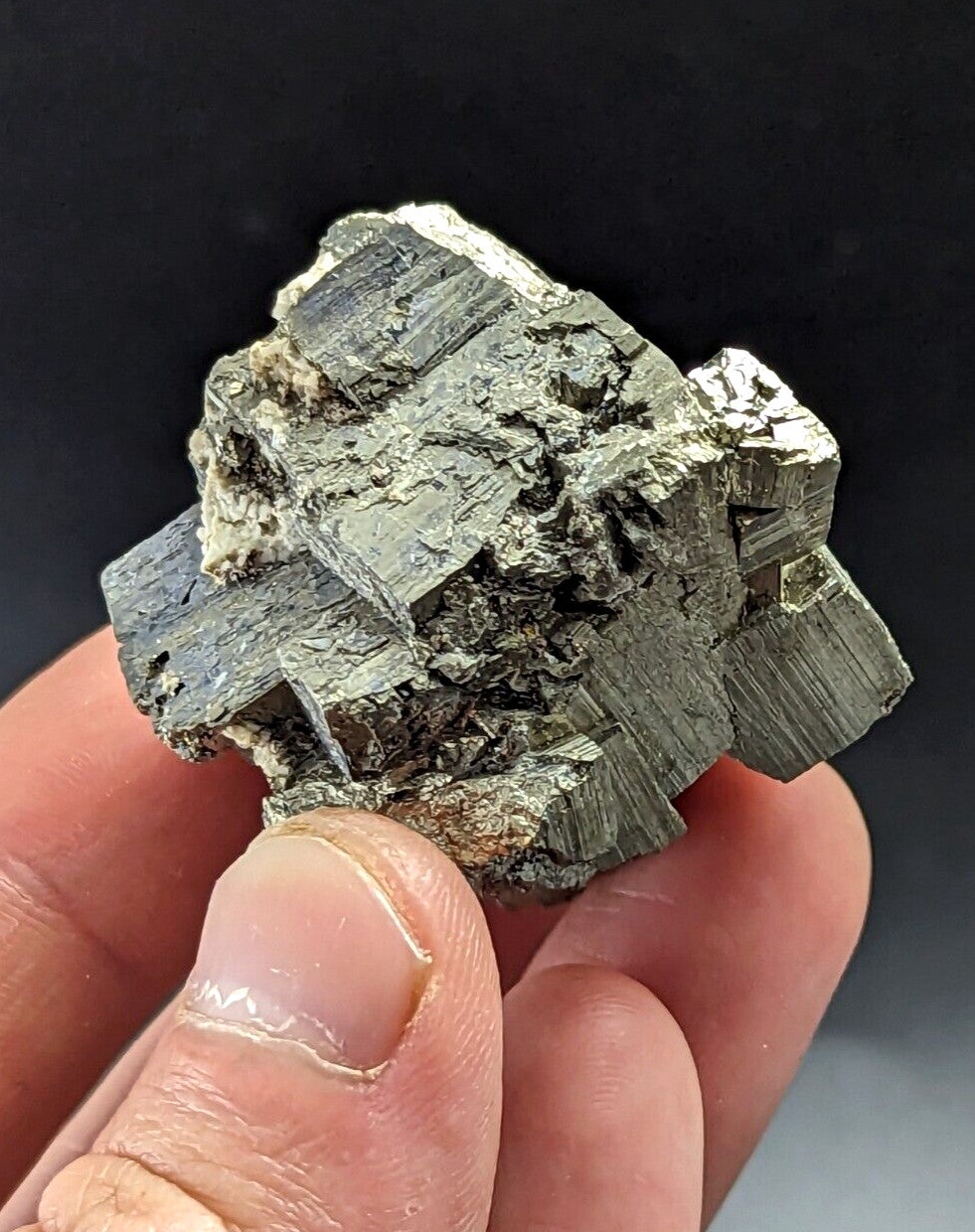 A hand holds a metallic, greenish-gray pyrite crystal cluster with distinct geometric facets and rough edges.