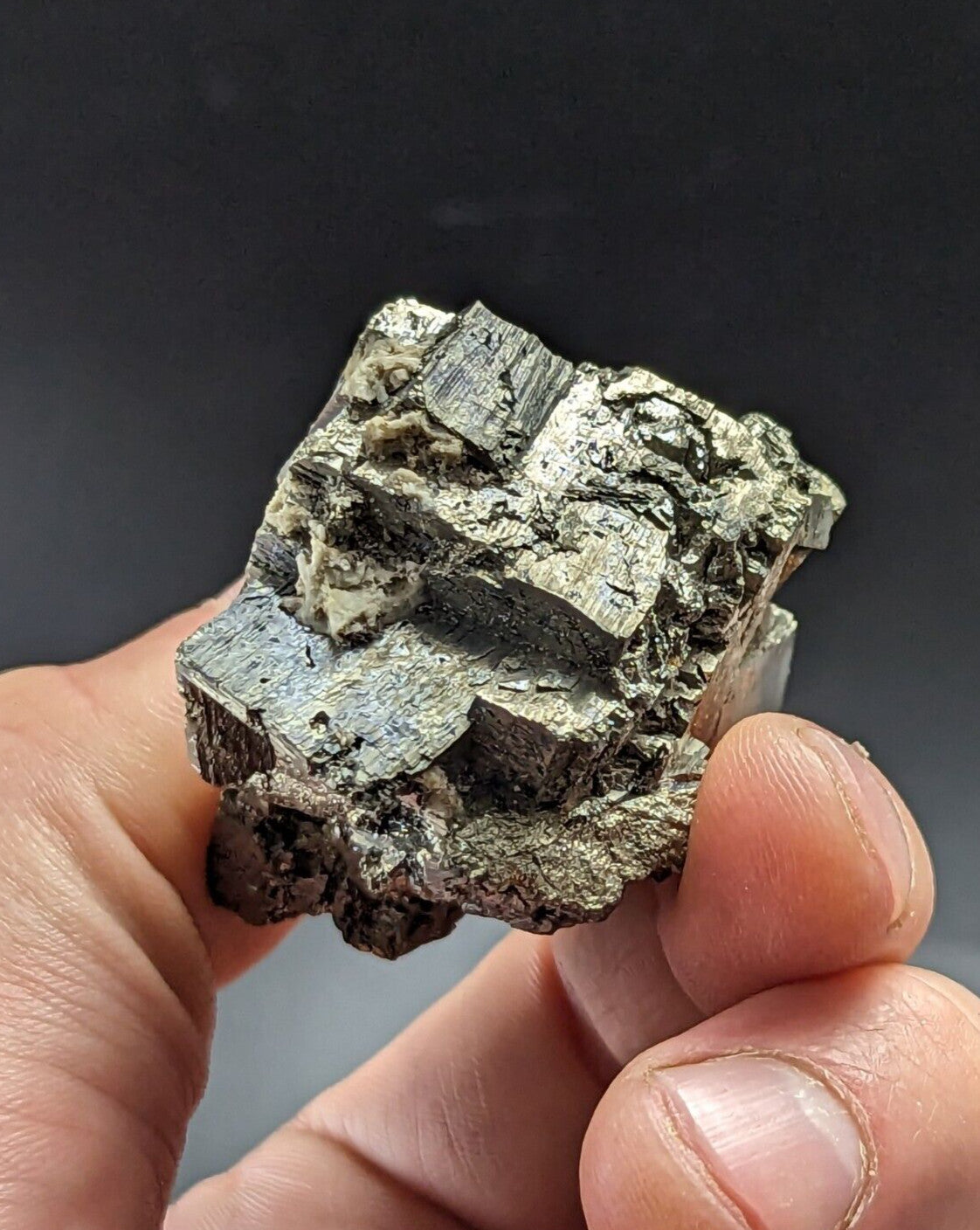 A hand holds a rough, metallic mineral specimen with distinct cubic crystals and a mottled greenish-gray surface.