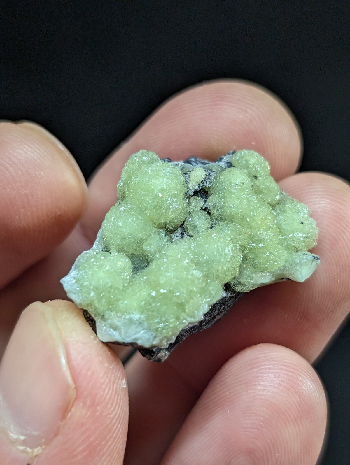 A cluster of translucent, pale green crystals with a rough, textured surface and dark matrix base.