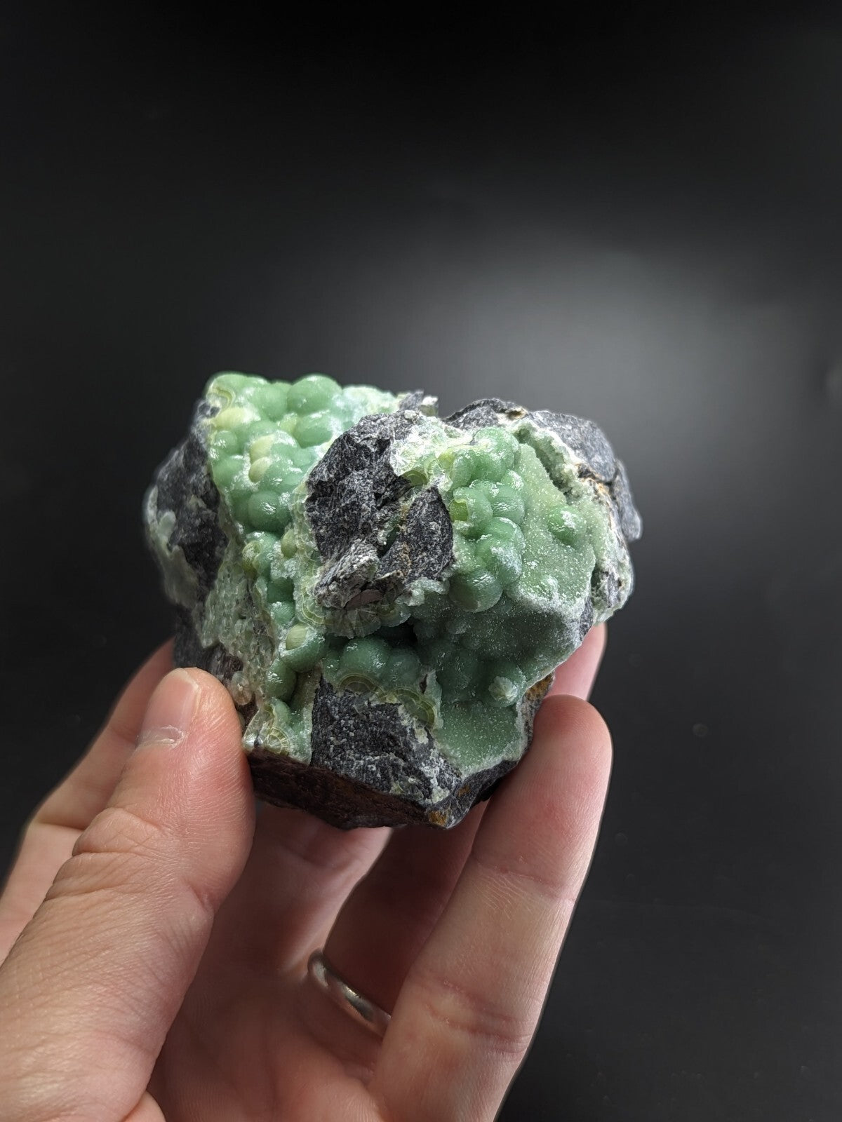 A hand holds a rough, irregularly shaped mineral specimen featuring vibrant green crystalline formations growing from dark gray rock.