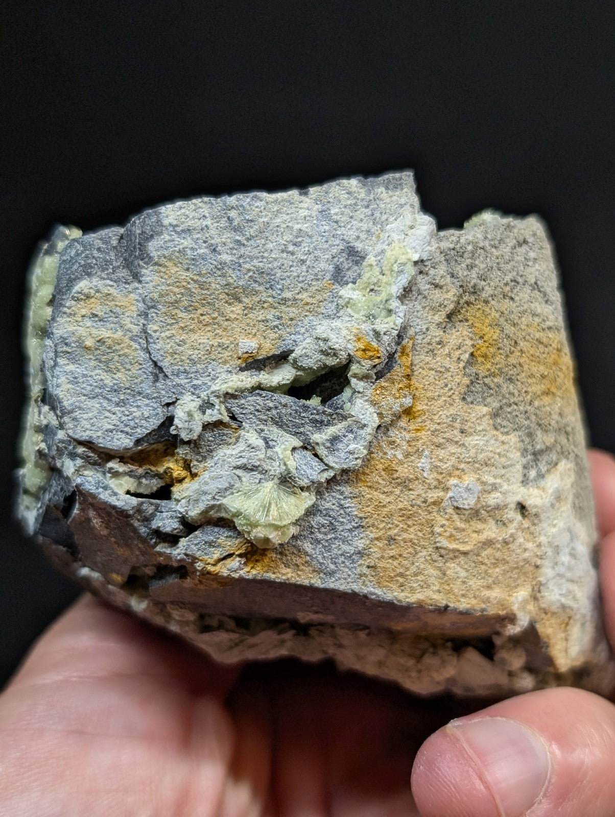 A rugged, irregularly shaped rock specimen with a mottled surface of gray, yellowish-orange, and pale green mineral deposits.