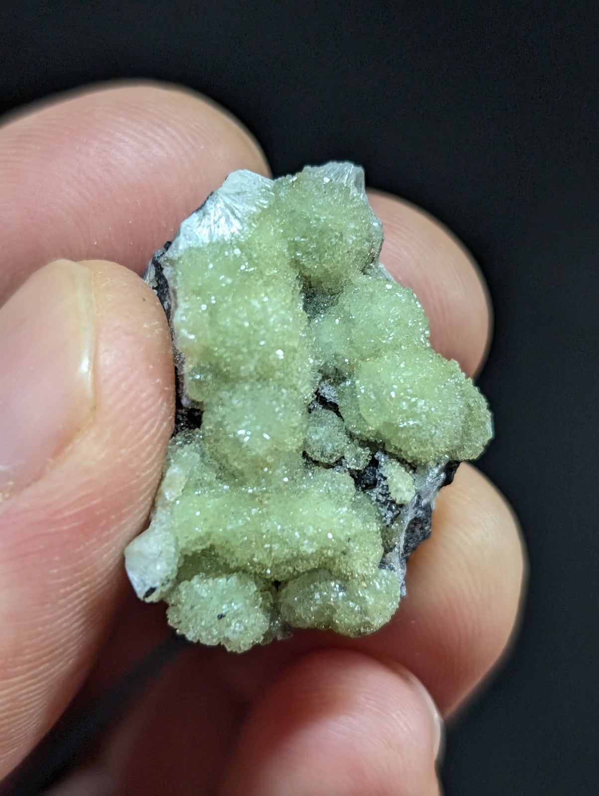 A pale green, crystalline mineral specimen with a rough, textured surface and embedded dark matrix.
