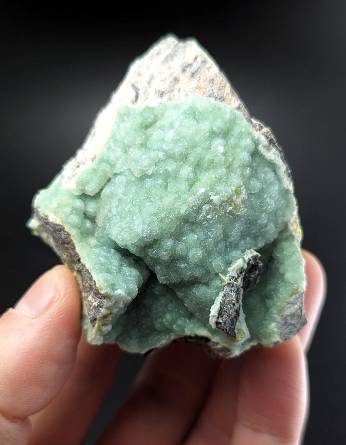 A hand holds a rough, light green mineral specimen with a textured surface of small crystalline formations against darker rock matrix.