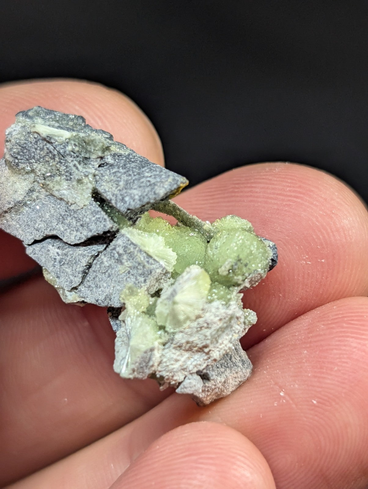 A hand holds a rough, gray rock specimen featuring clusters of translucent green crystals.