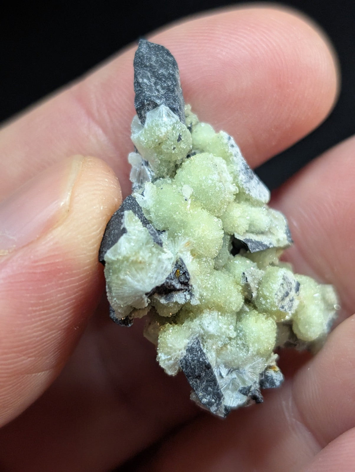 A cluster of pale green, crystalline minerals with dark gray rock matrix.