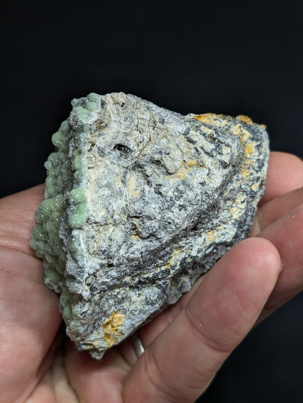 A hand holds a rough, irregularly shaped mineral specimen featuring patches of pale green crystalline growths against a grayish matrix with speckles of yellow and black.