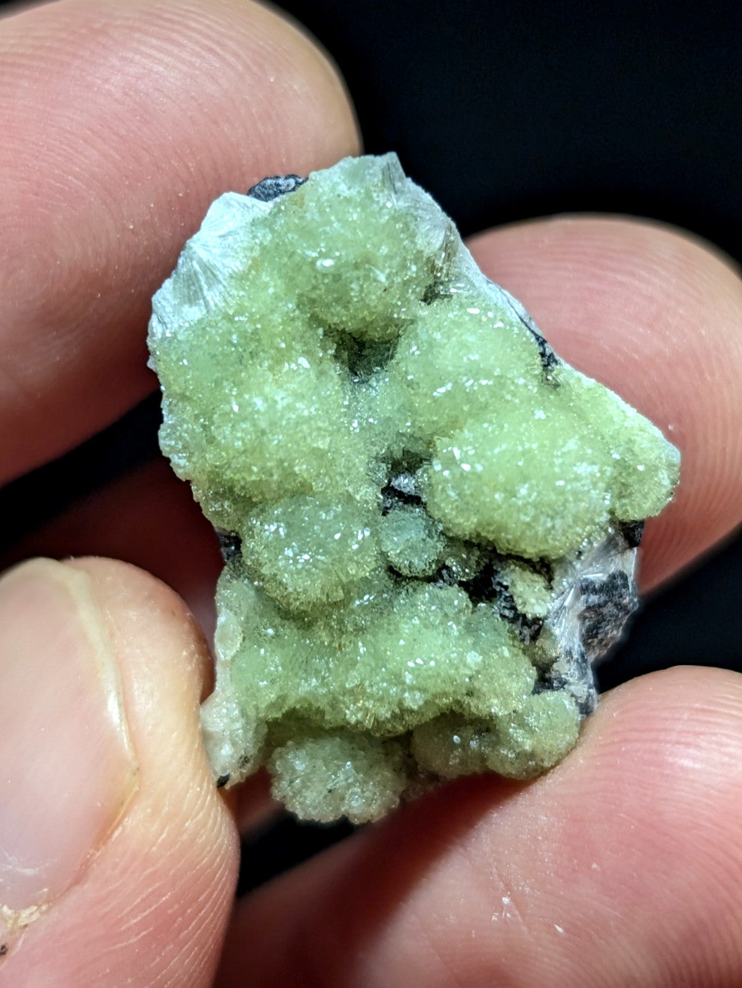 A pale green, crystalline mineral specimen with a rough, textured surface and embedded darker matrix.