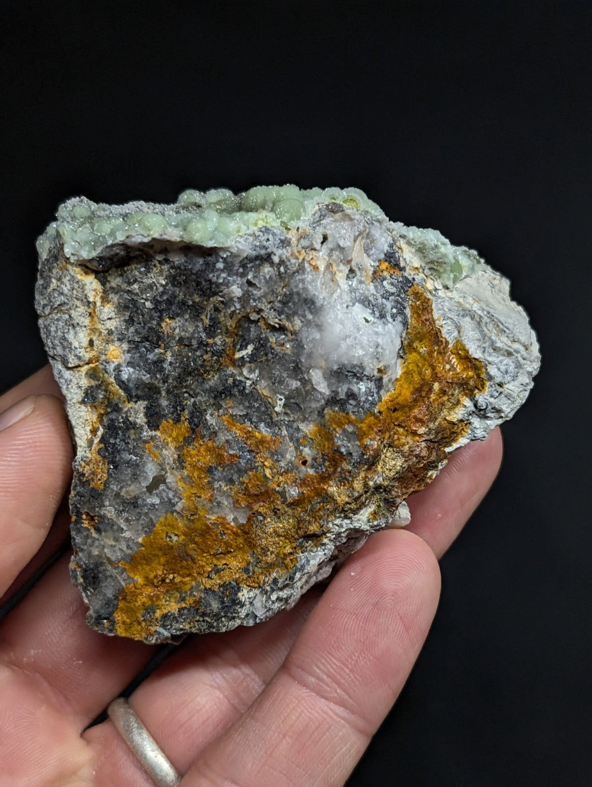 A hand holds a rough, irregularly shaped mineral specimen featuring patches of gray rock with vibrant orange and pale green crystalline formations.