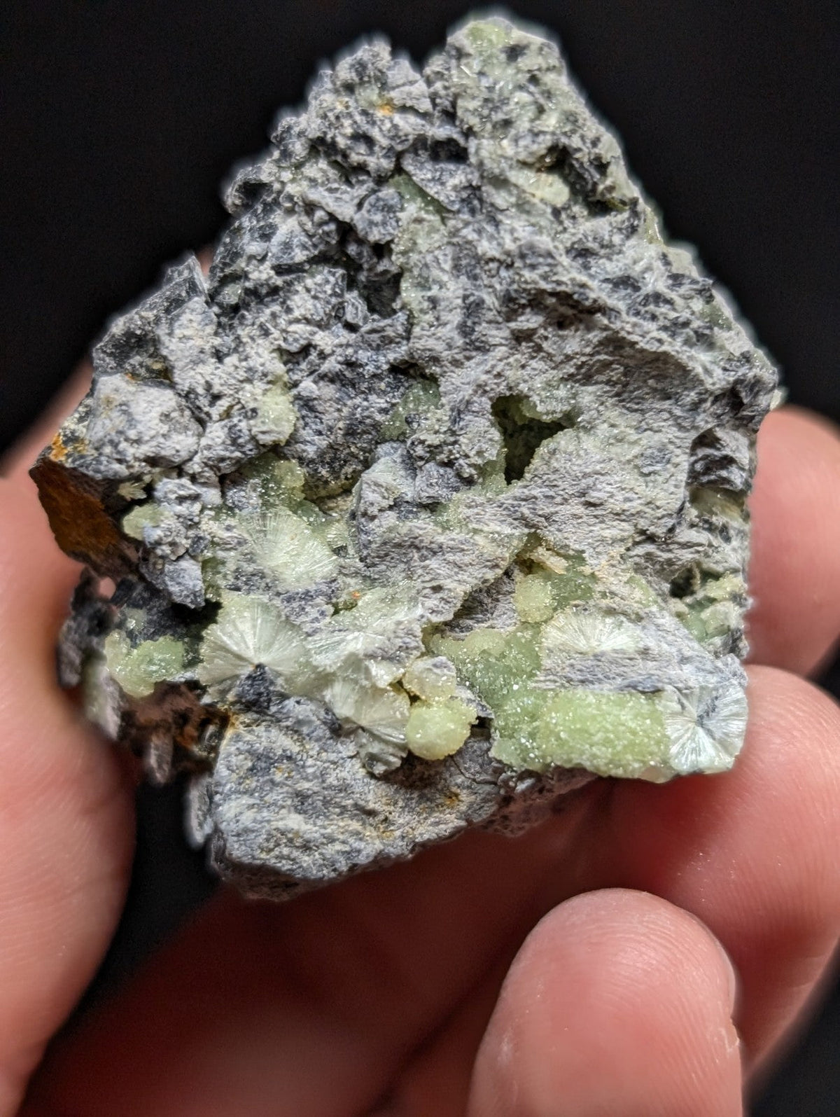 A hand holds a rough, irregularly shaped mineral specimen featuring grayish matrix with prominent pale green crystalline formations.